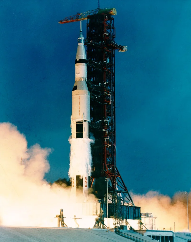 Apollo 11’s Saturn V rising beside the launch tower, with bright exhaust and smoke below.