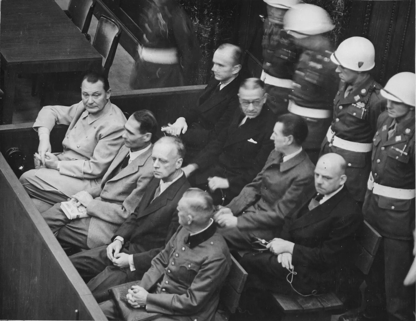 Eight defendants seated in two rows in the Nuremberg courtroom, with uniformed military guards behind them.