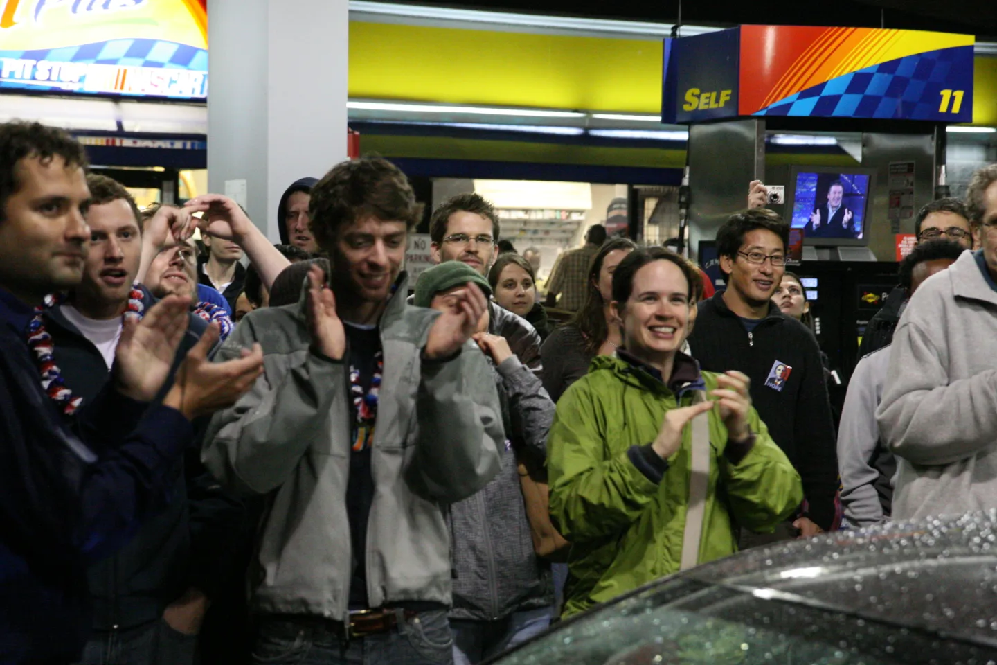 People applauding beneath a gas-station canopy, with a small television behind the group.