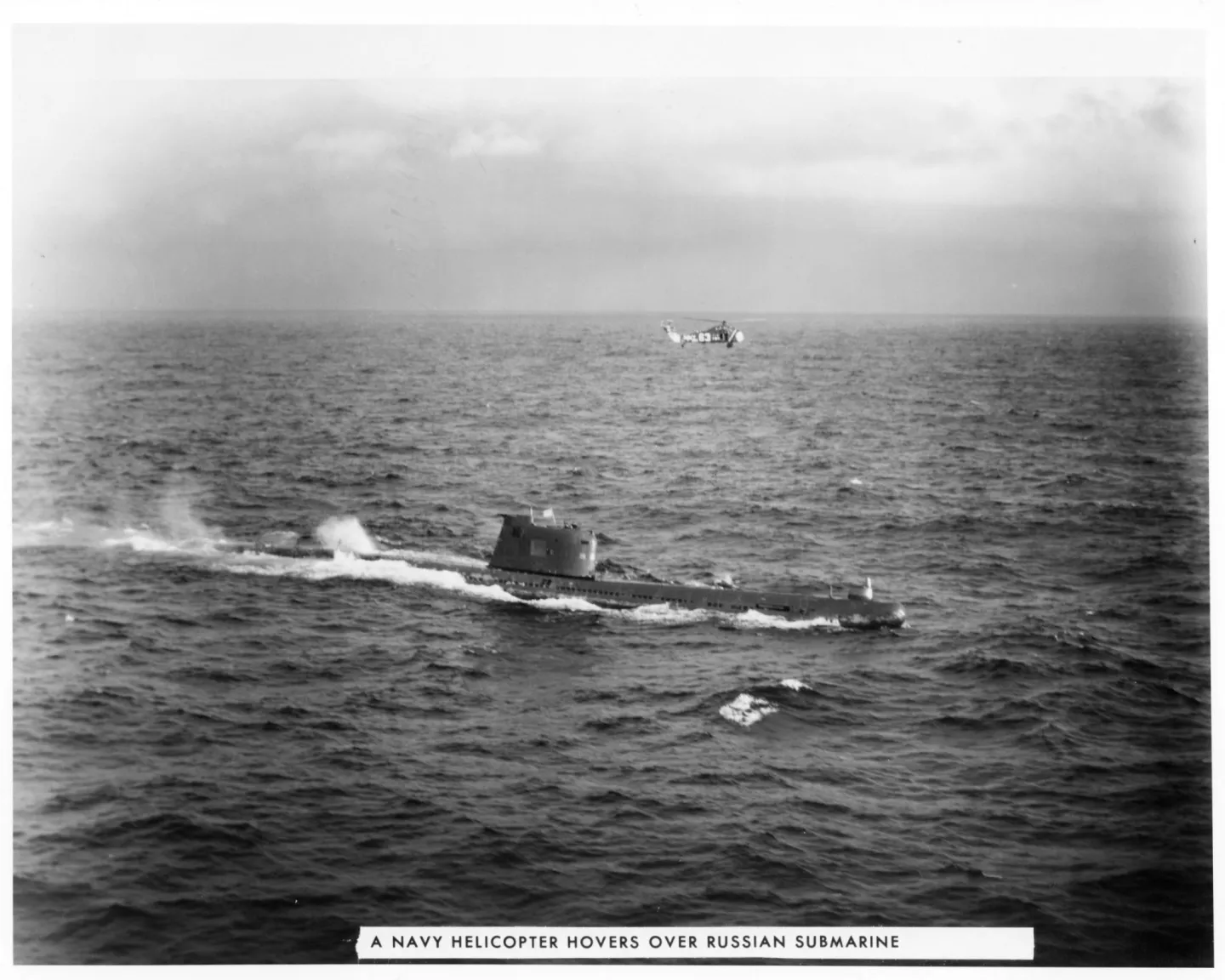 A surfaced Soviet submarine underway at sea with a U.S. Navy helicopter hovering beyond it during the Cuban quarantine.