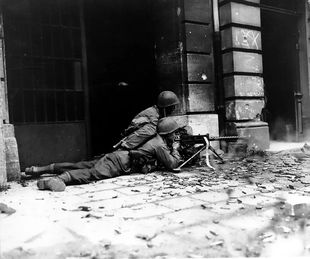 Two American infantrymen at a building entrance with a Browning machine gun aimed along an Aachen street.