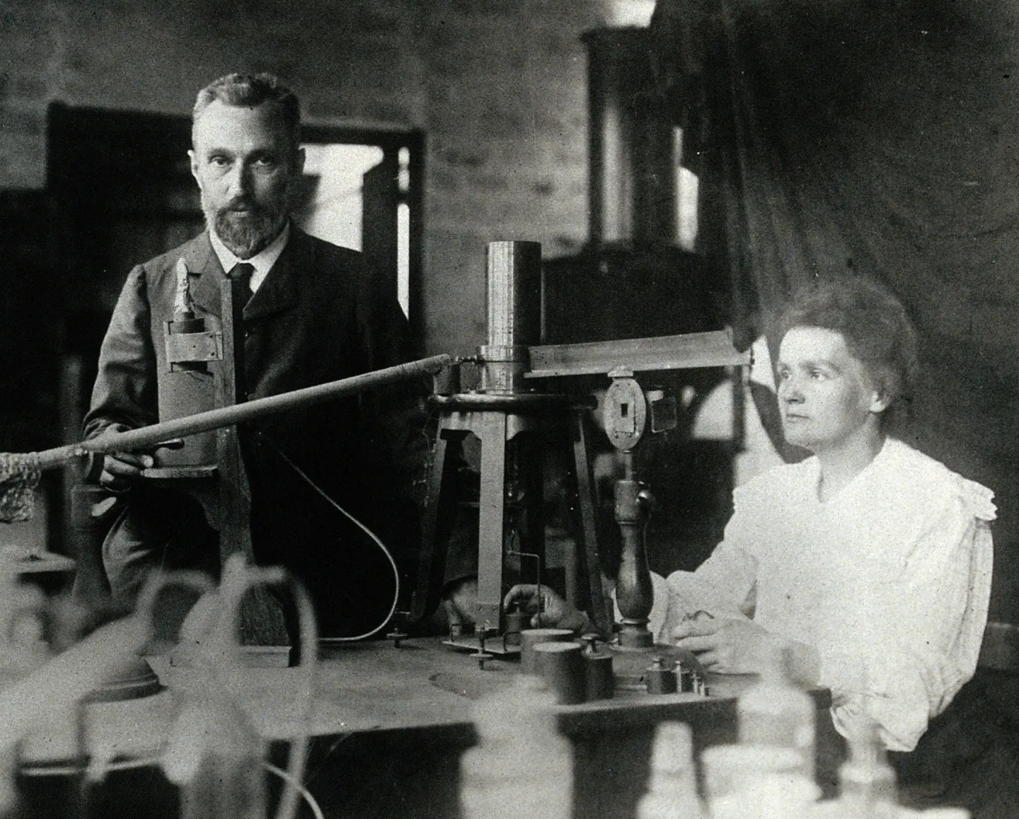 Pierre Curie sits at left while Marie Curie works beside laboratory measuring equipment.