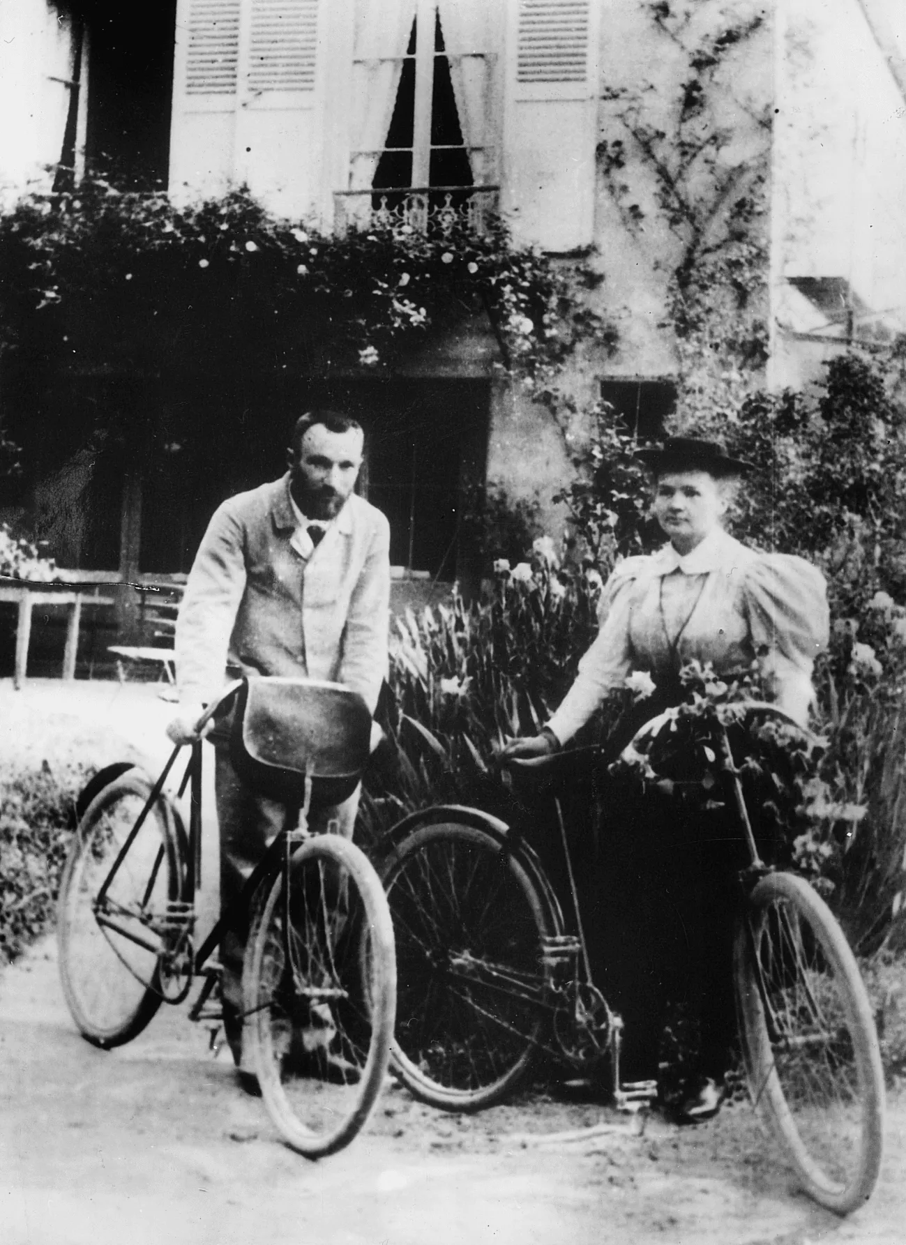 Pierre and Marie Curie stand outdoors beside two bicycles.