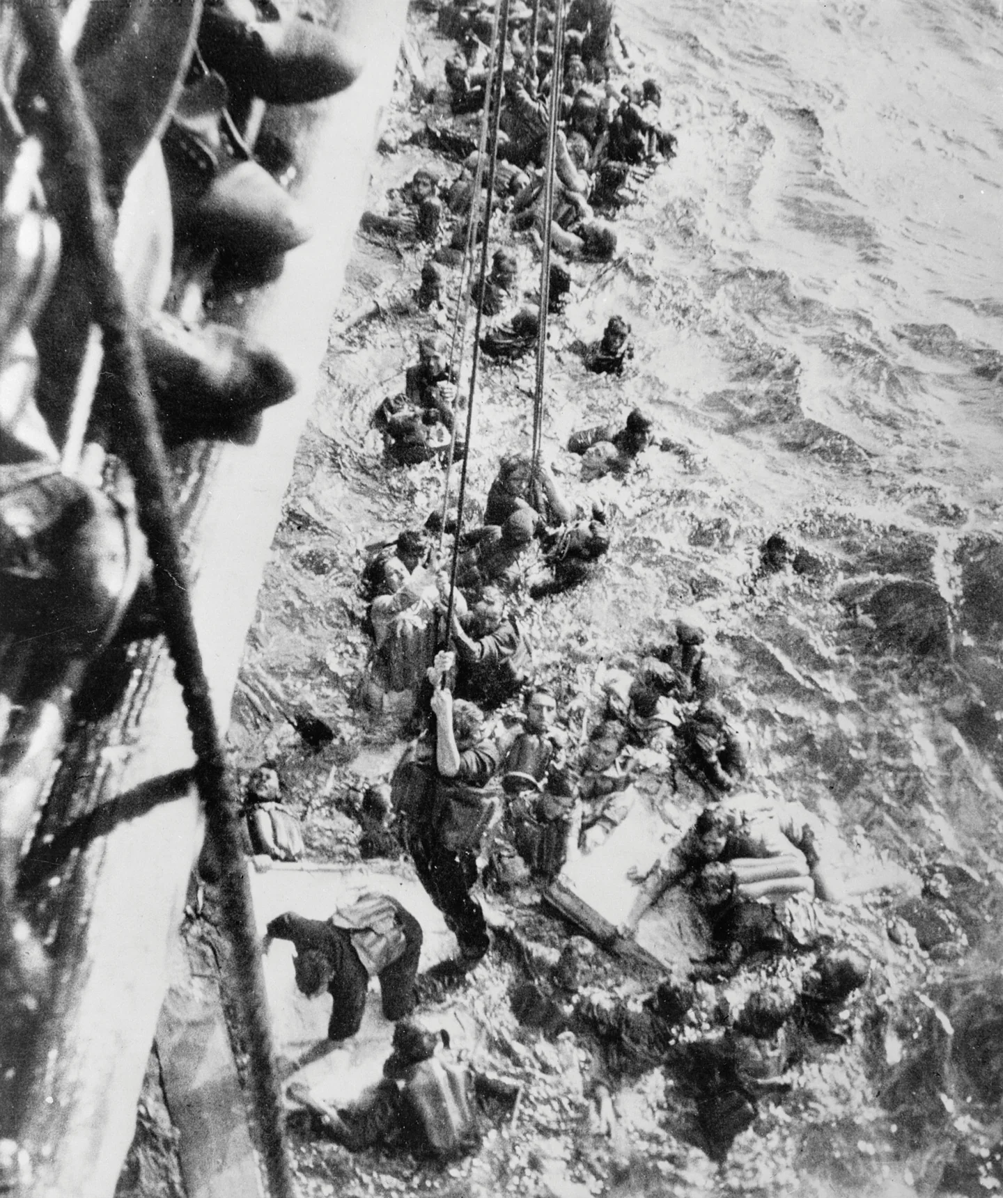 Bismarck survivors in the sea beside HMS Dorsetshire, with ropes lowered down the ship’s side to help men reach safety.