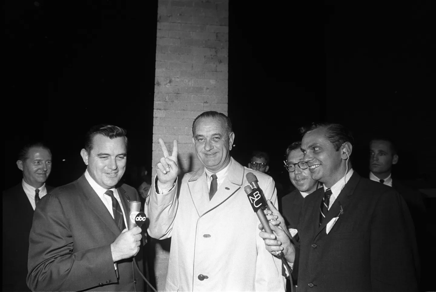 Johnson raises two fingers in a victory sign while reporters hold microphones beside him.
