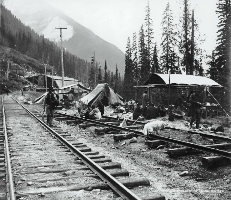 Workers, tents and equipment sit beside railway tracks in a mountain valley.