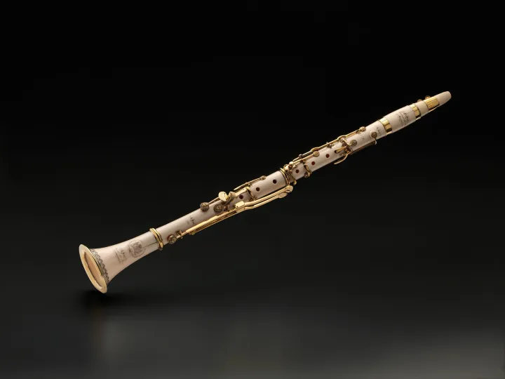 White ivory clarinet with gold-colored keys lying diagonally against a dark background.