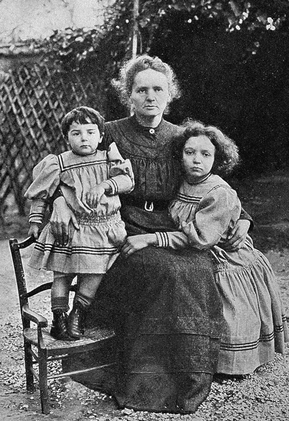 Marie Curie sits outdoors with her two daughters beside her.