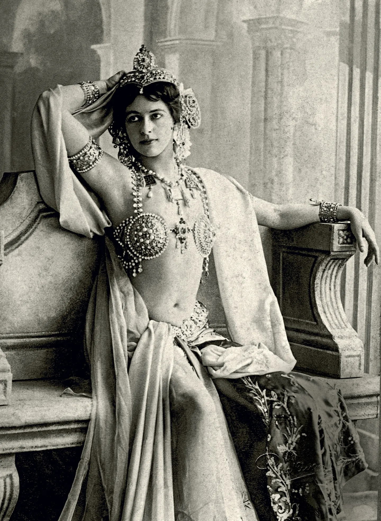 Mata Hari seated in an ornate stage costume, with one arm raised to her jeweled headdress.