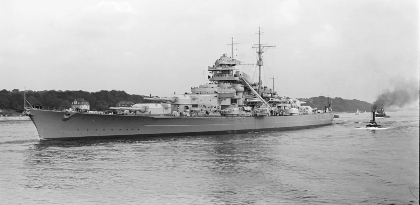 Black-and-white 1940 photograph of Bismarck off Blankenese, with forward gun turrets, a wooded shore and tugs near the stern.