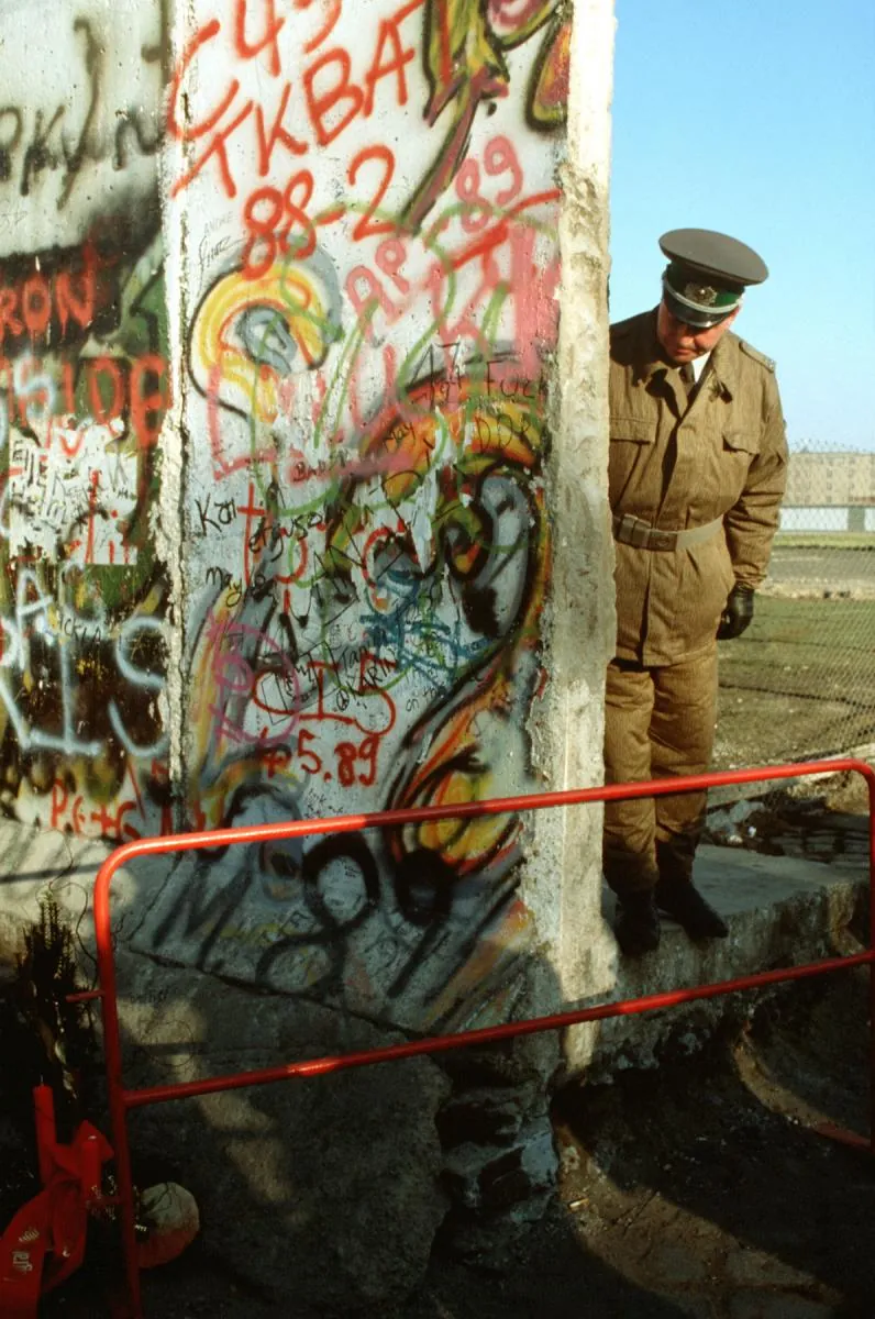 An East German policeman looking around a broken, graffiti-covered section of the Berlin Wall.