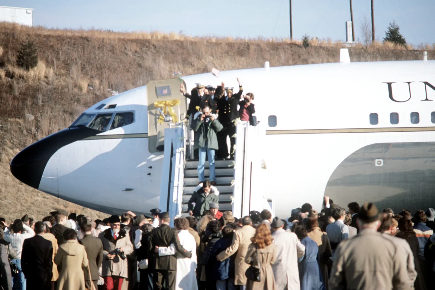 Former hostages waving from an aircraft doorway above waiting families and photographers.