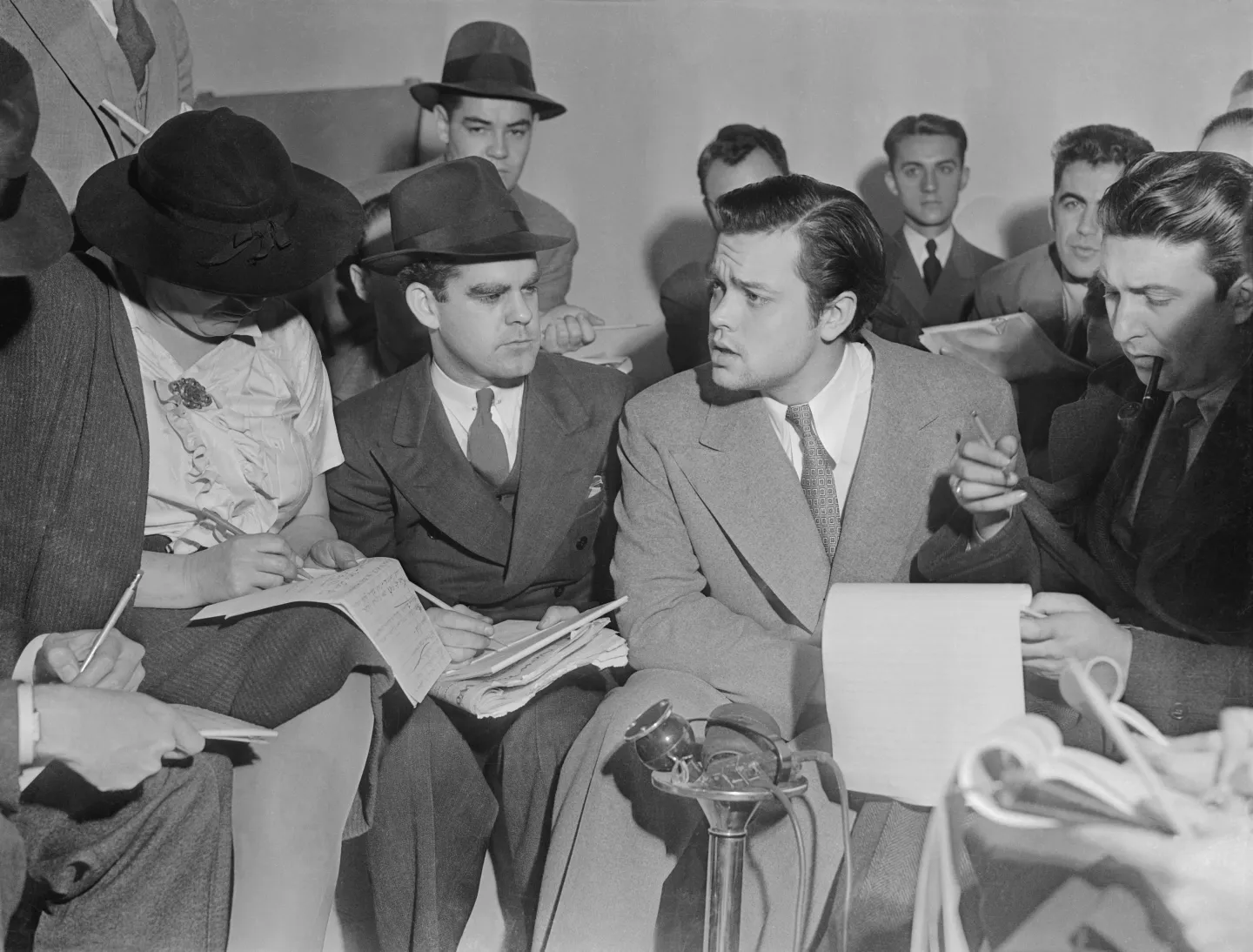 Orson Welles surrounded by reporters and microphones at a press conference.