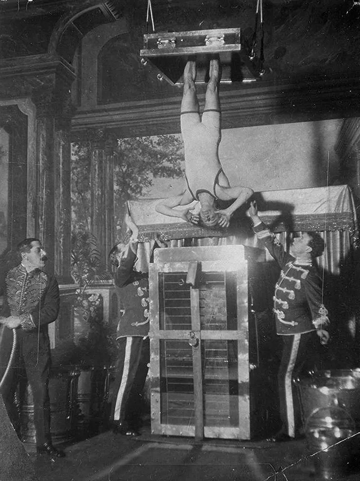 Assistants hold Houdini upside down above the water torture cell.