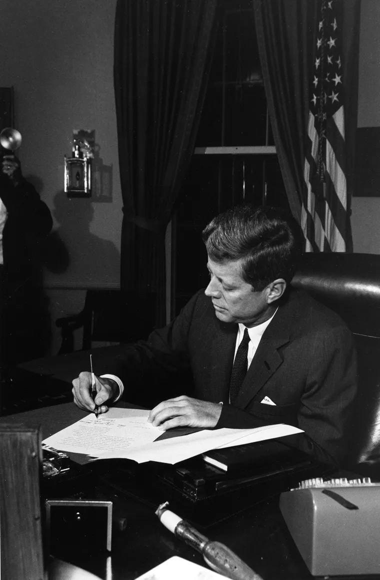 President John F. Kennedy signing the Cuba quarantine proclamation at his Oval Office desk, with the American flag behind him.
