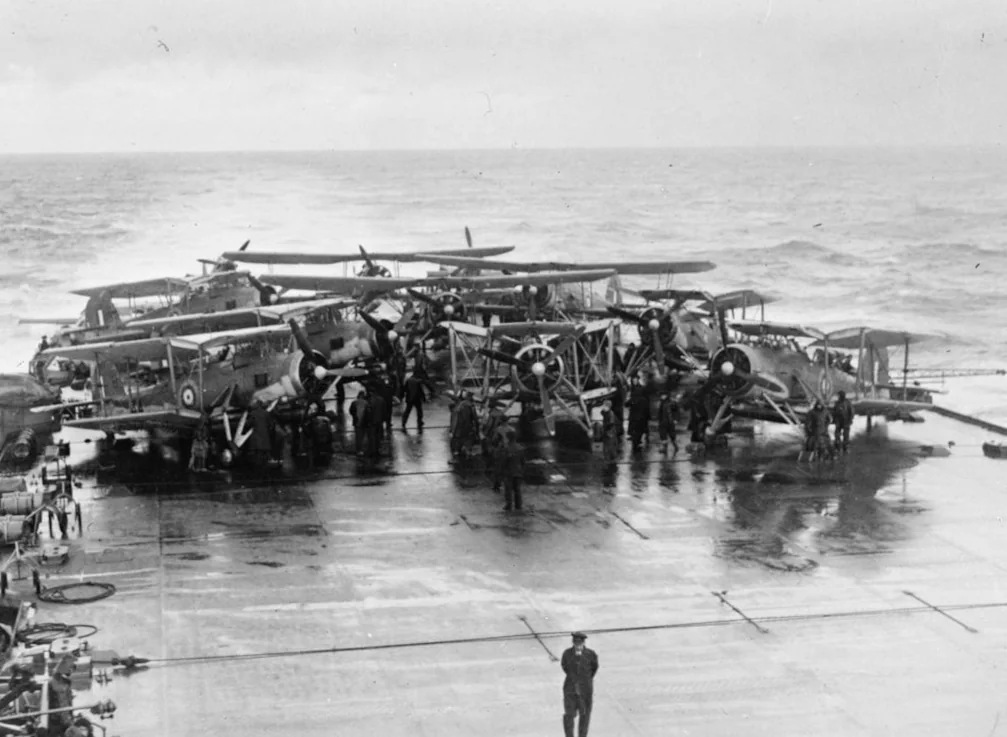 Swordfish biplanes with folded wings and crews crowded together on the wet flight deck of HMS Victorious.
