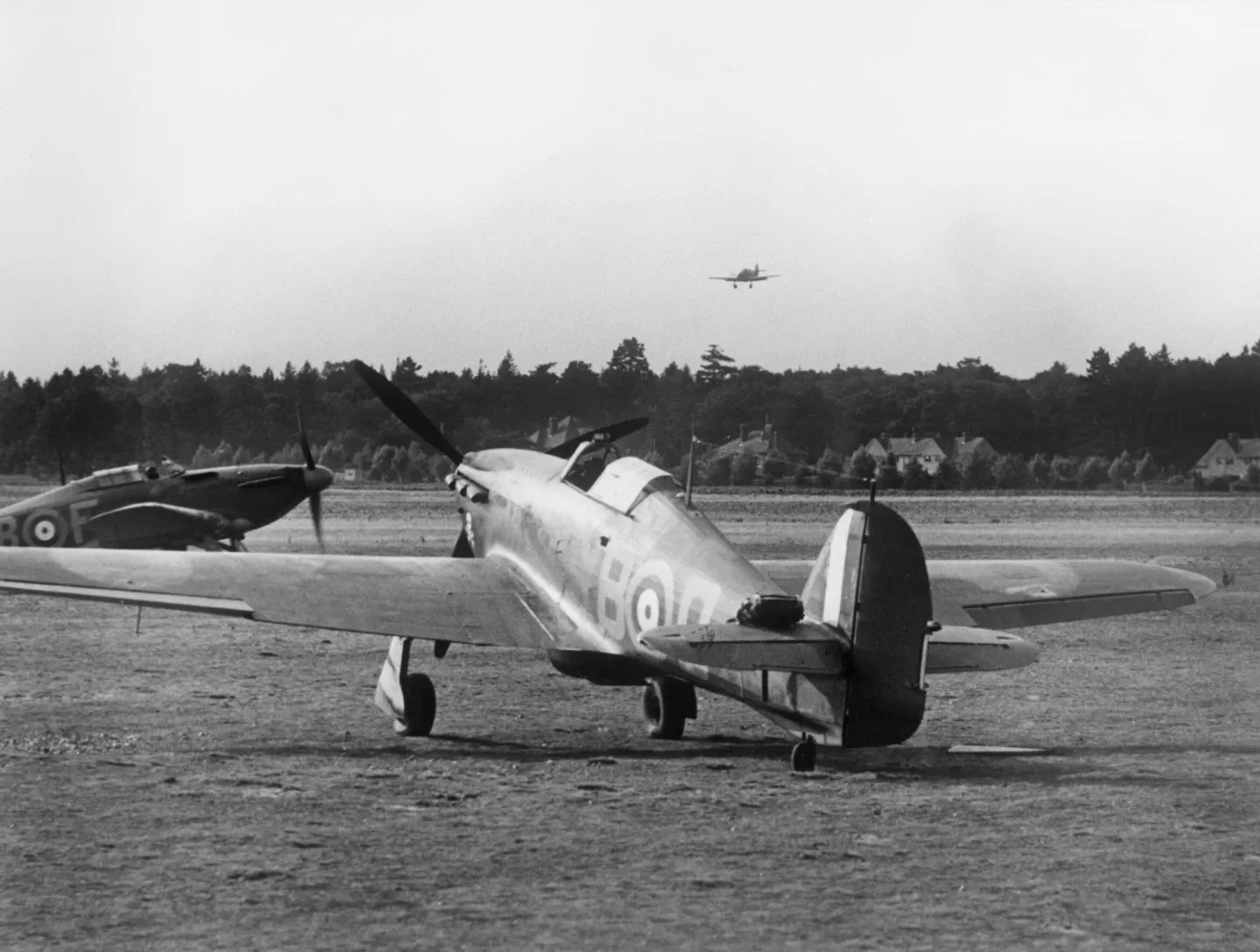 Hurricanes parked on a grass airfield while another flies low above them.