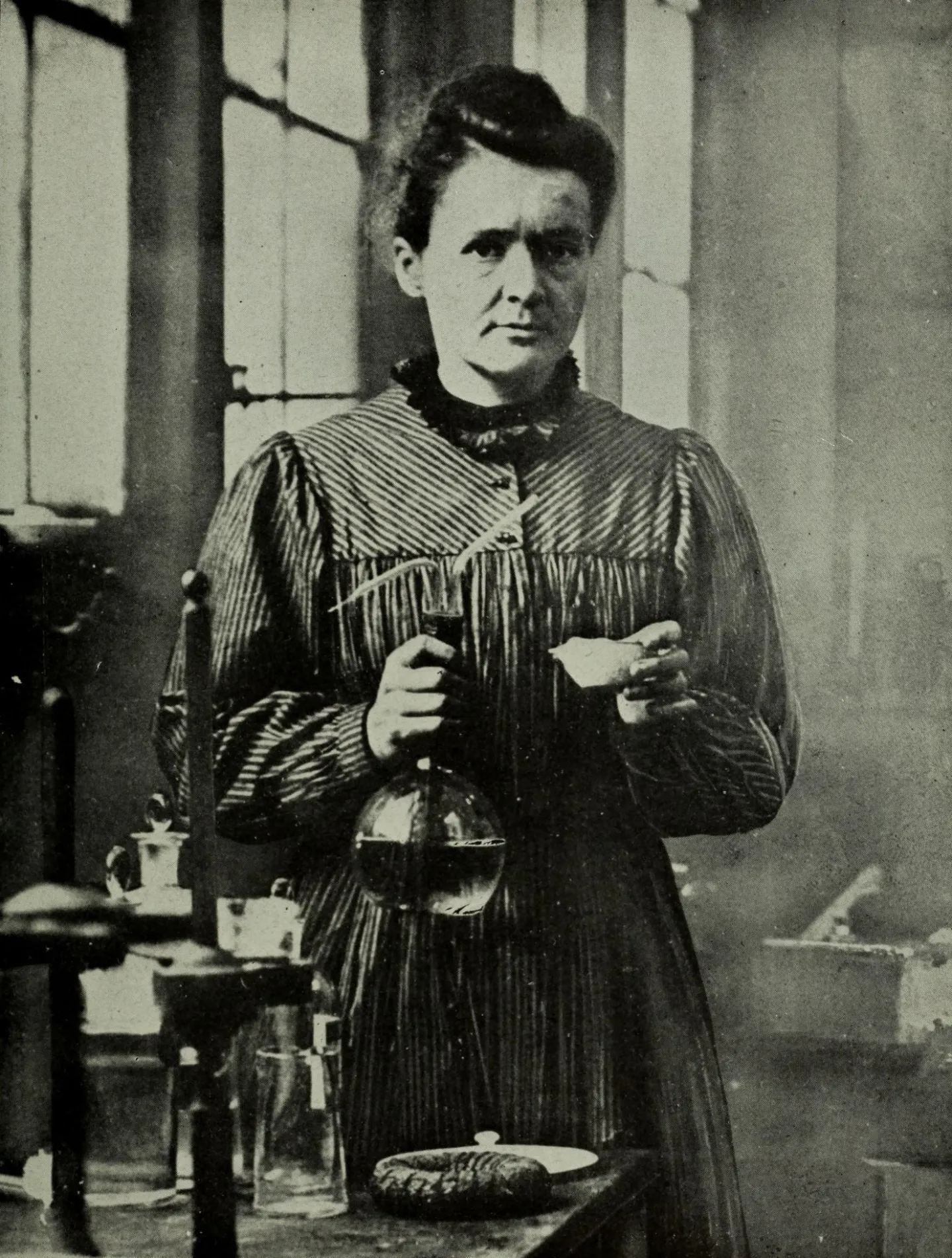 Marie Curie stands beside laboratory bottles holding a glass vessel.