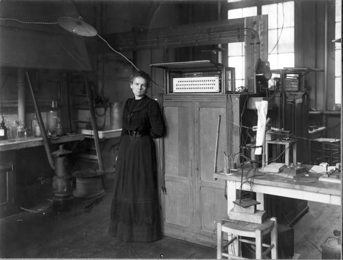 Marie Curie stands between benches and cabinets in a large laboratory.