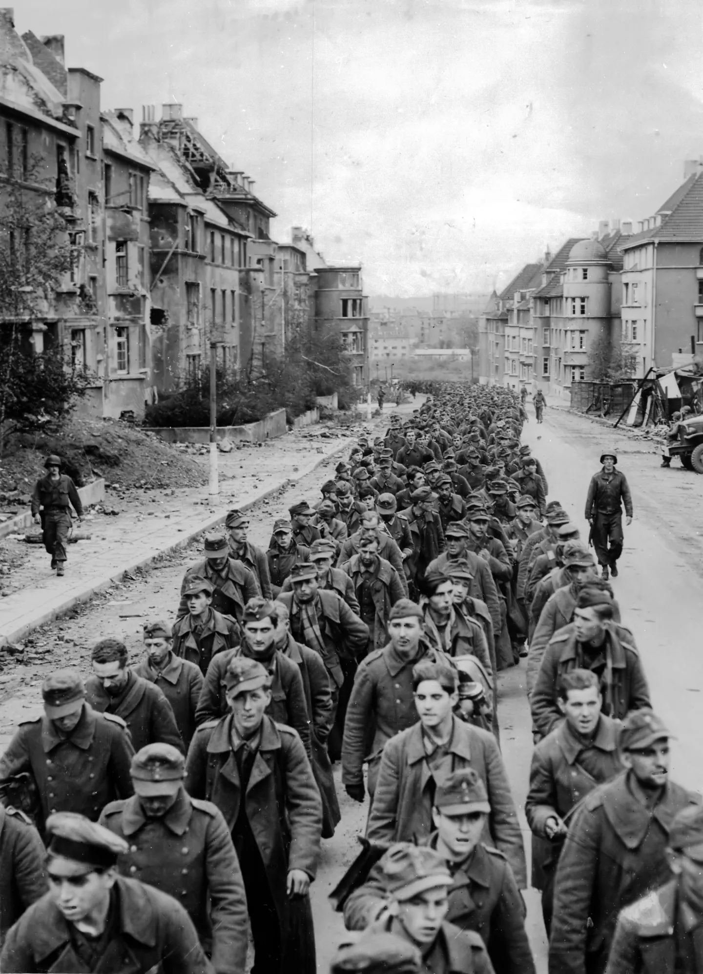 A long column of German prisoners marching through ruined streets in Aachen, watched by American guards.