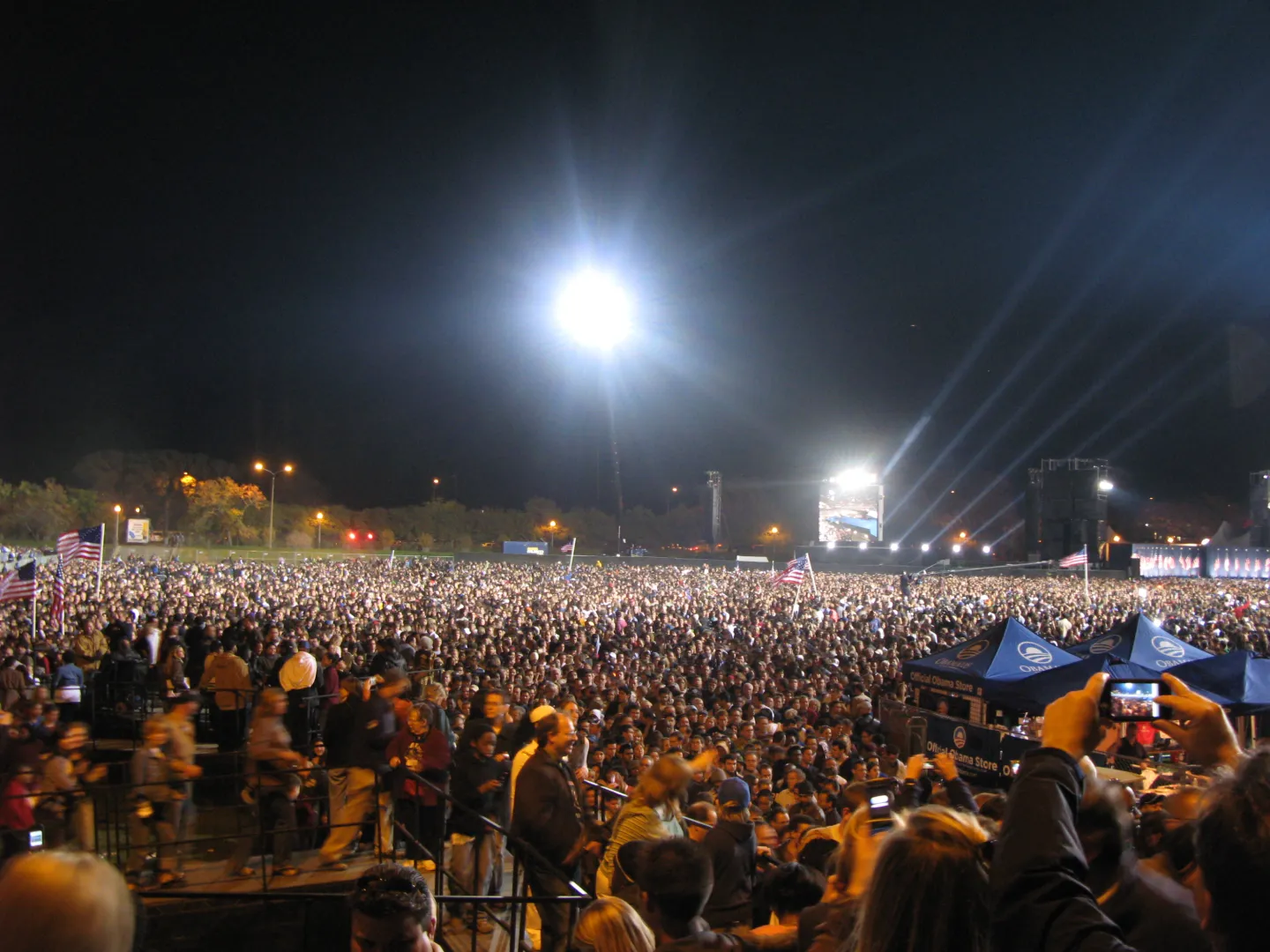A dense nighttime crowd in Grant Park under floodlights, with flags and a distant stage.