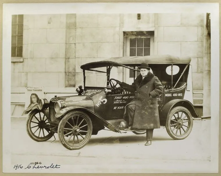 A person stands beside a touring car cataloged as a 1916 Chevrolet.