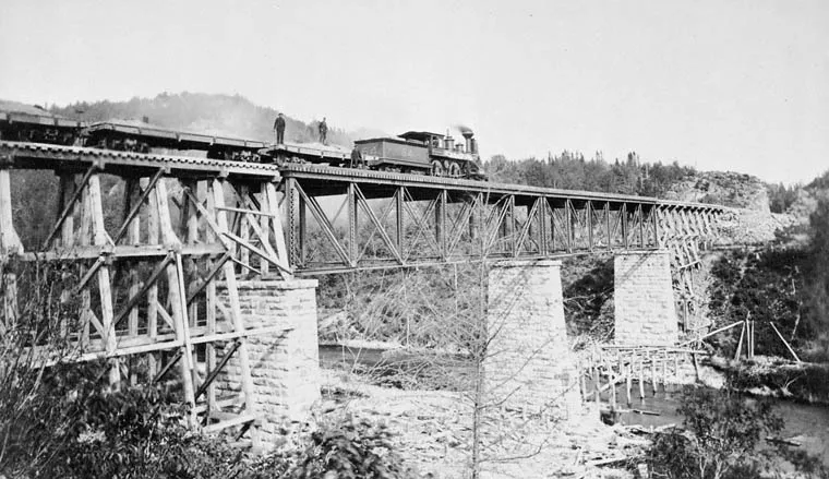 A steam locomotive and cars stand on a high bridge over a rocky river valley.