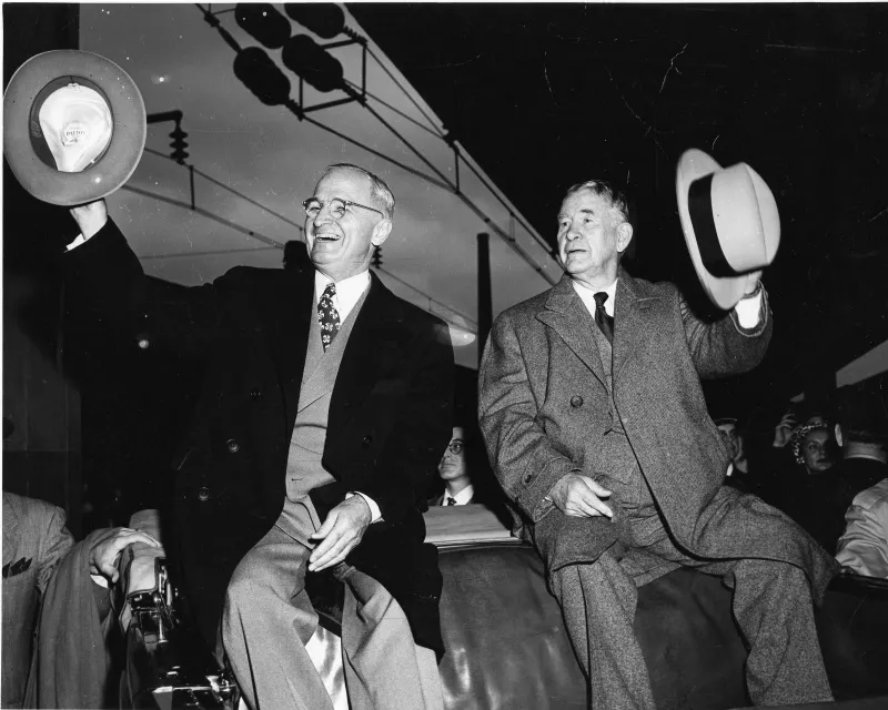 Truman and Alben Barkley seated in an open vehicle, waving their hats.