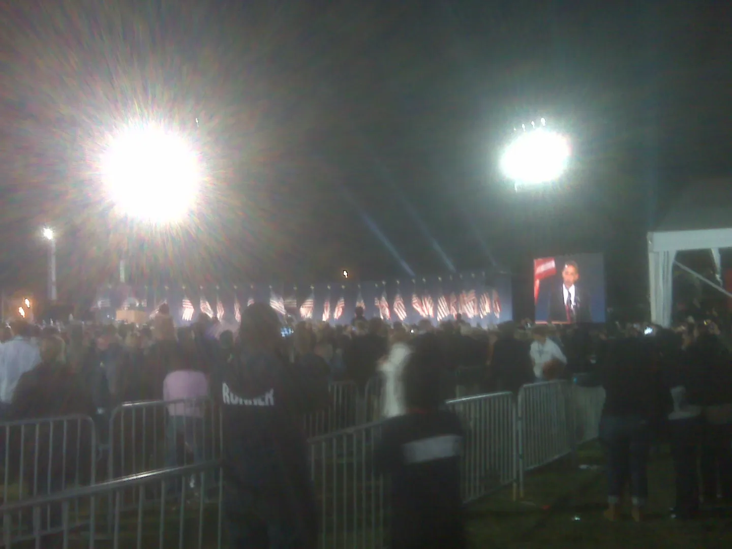 A distant election-night stage and large screen showing Obama at a lectern, photographed over spectators.