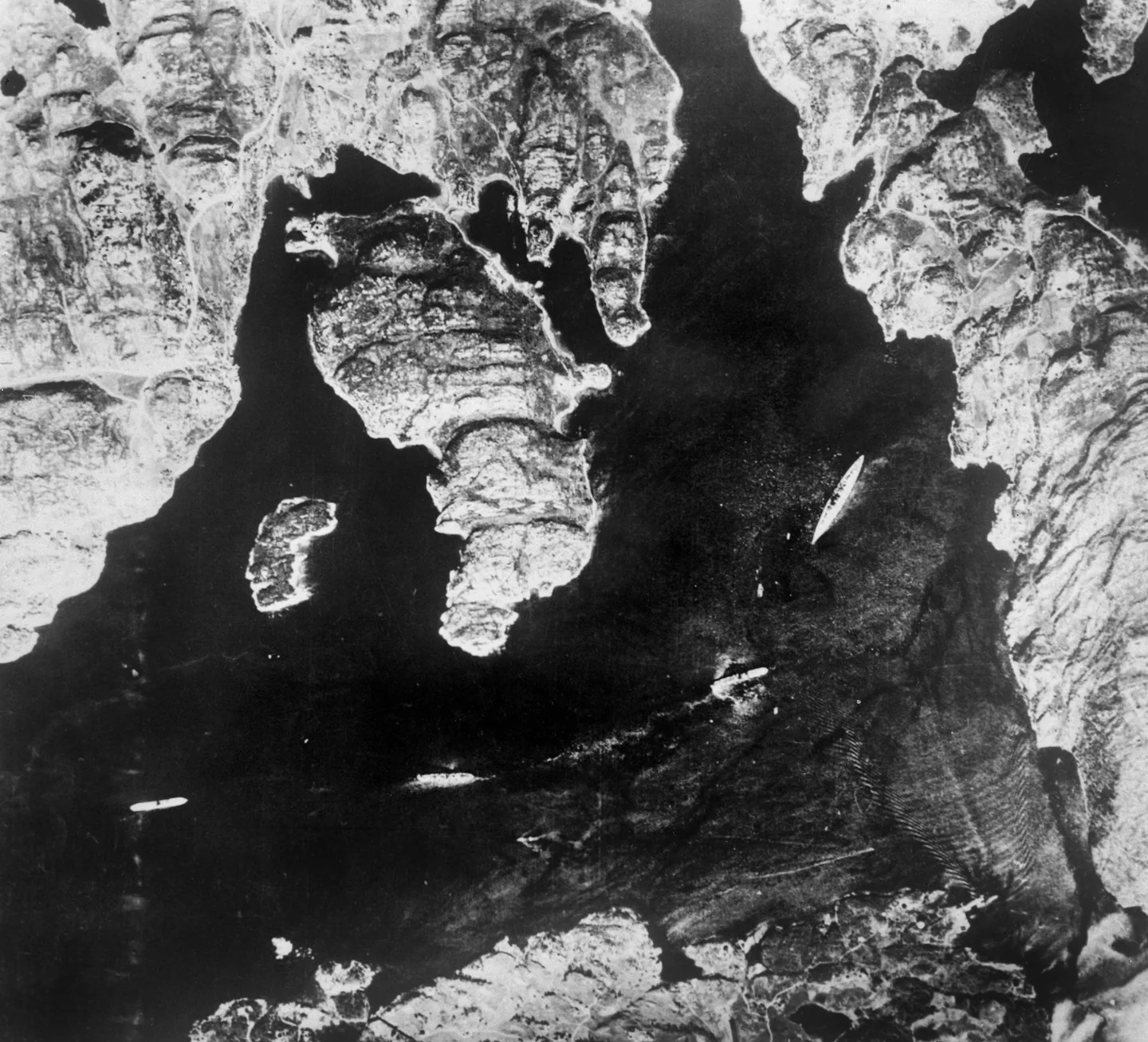 Aerial reconnaissance photograph of Bismarck as a pale ship in a dark Norwegian fjord, surrounded by an irregular rocky coastline.