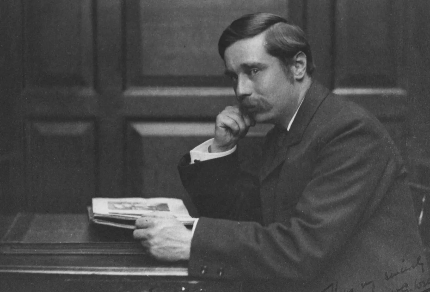 H. G. Wells seated at a table with a book and one hand against his face.