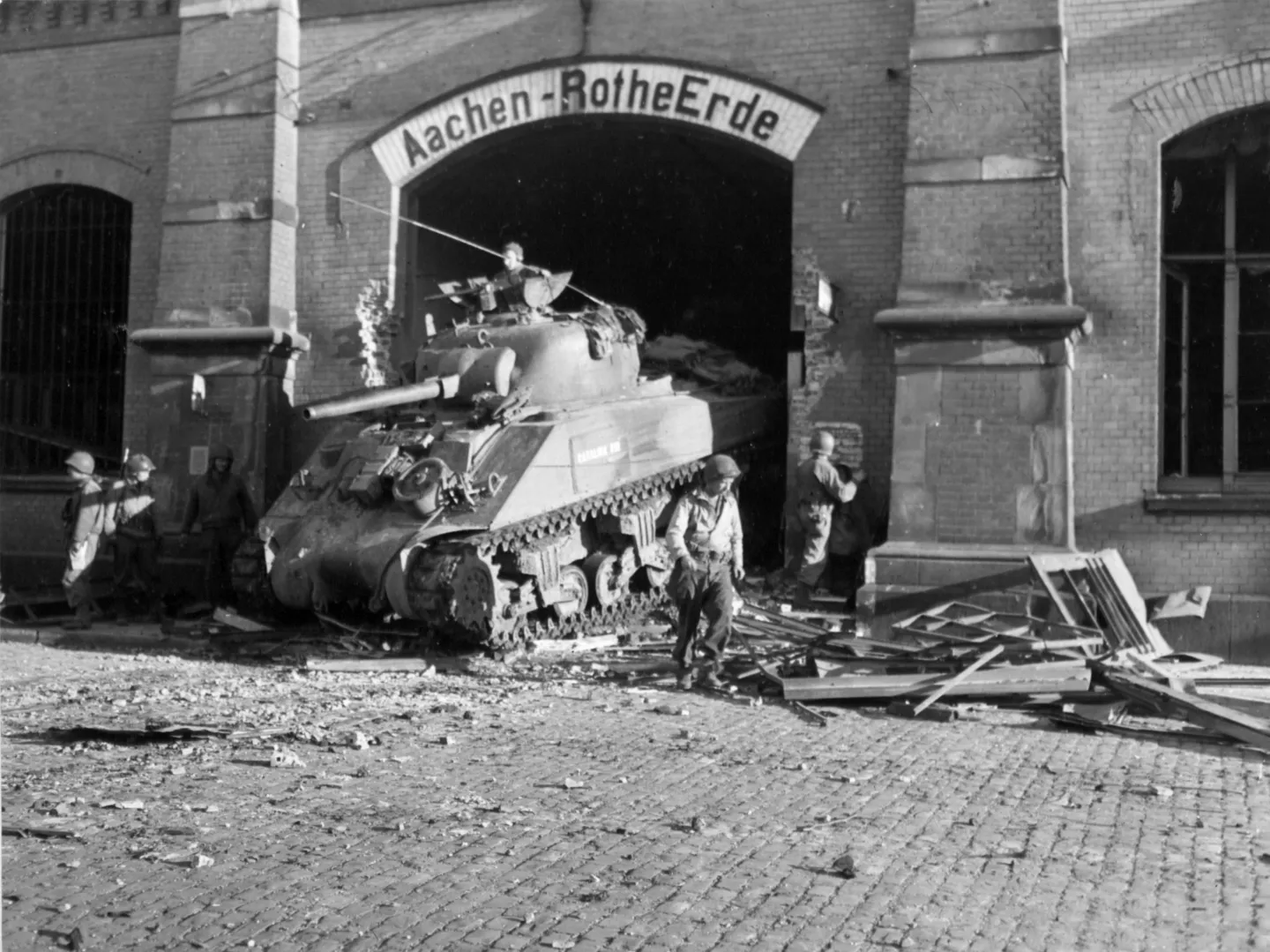 An M4 Sherman tank emerging through an opening beneath the Aachen-Rothe Erde railway station sign.