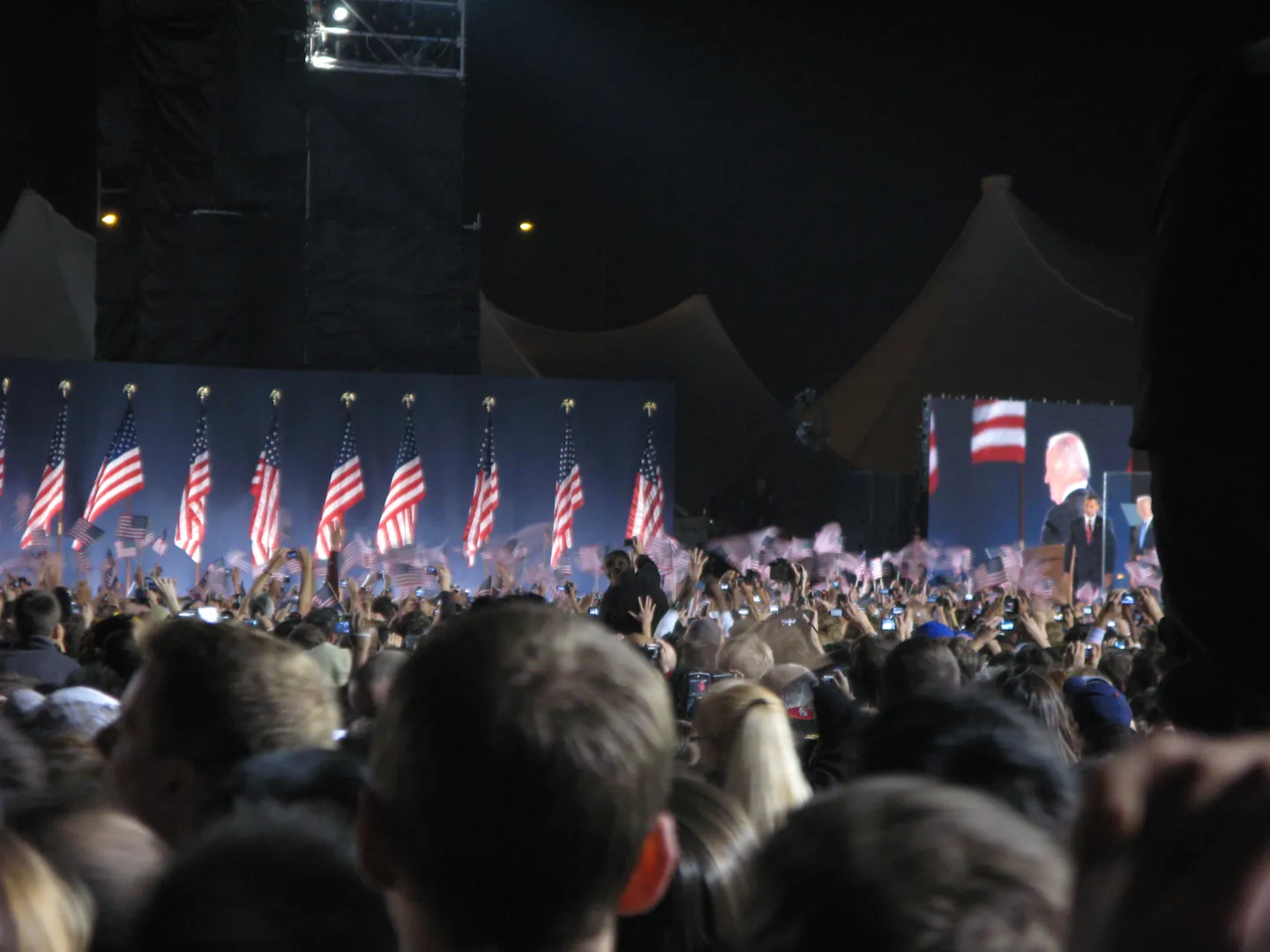Joe Biden displayed on a large screen while the crowd raises cameras and flags.