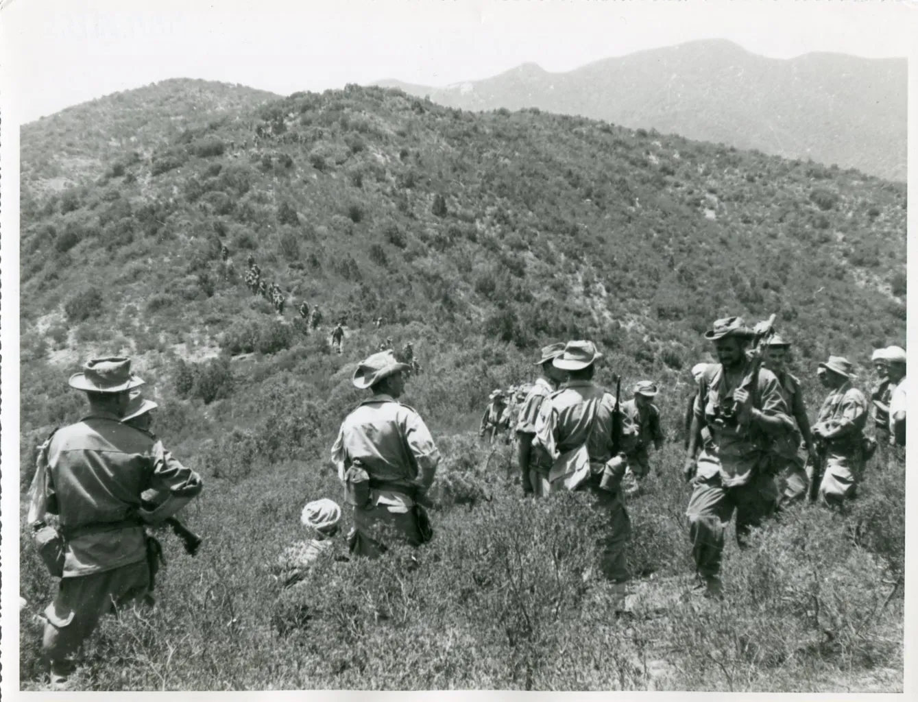 Armed Harkis walk in a line along a mountain ridge.