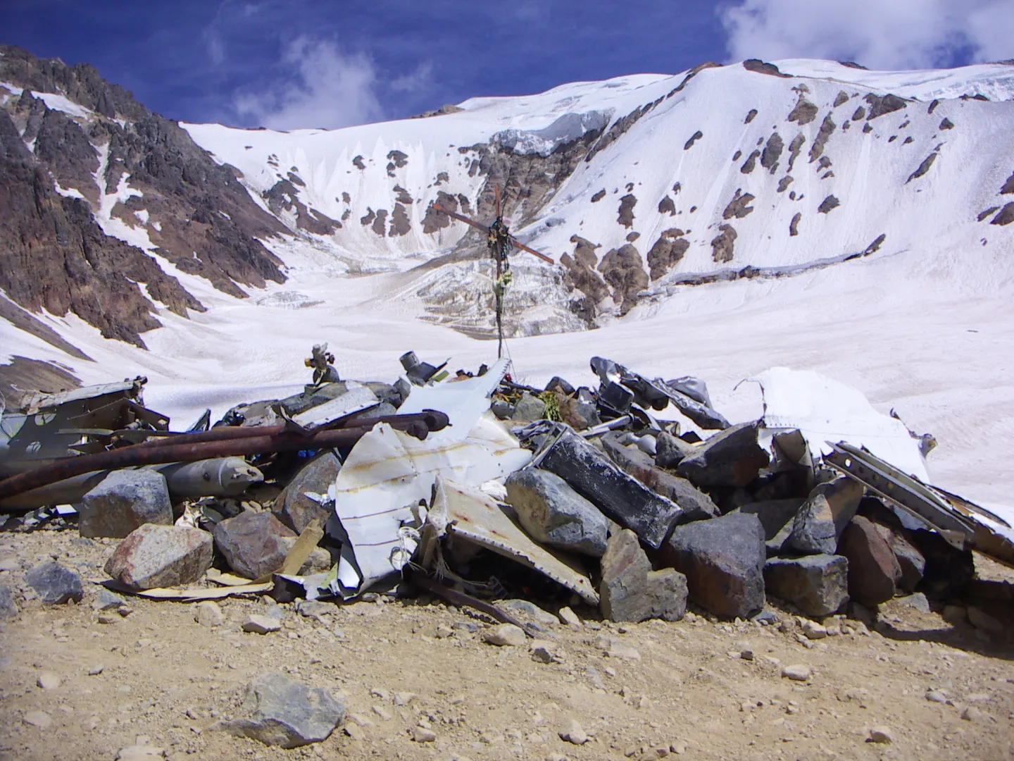 A cross and scattered aircraft fragments at the Flight 571 memorial below steep, snow-covered mountains.