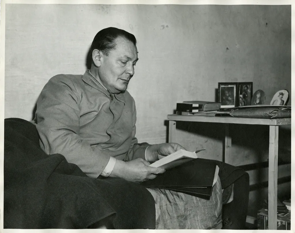 Hermann Göring sits reading in his prison cell beside a table holding framed family photographs.