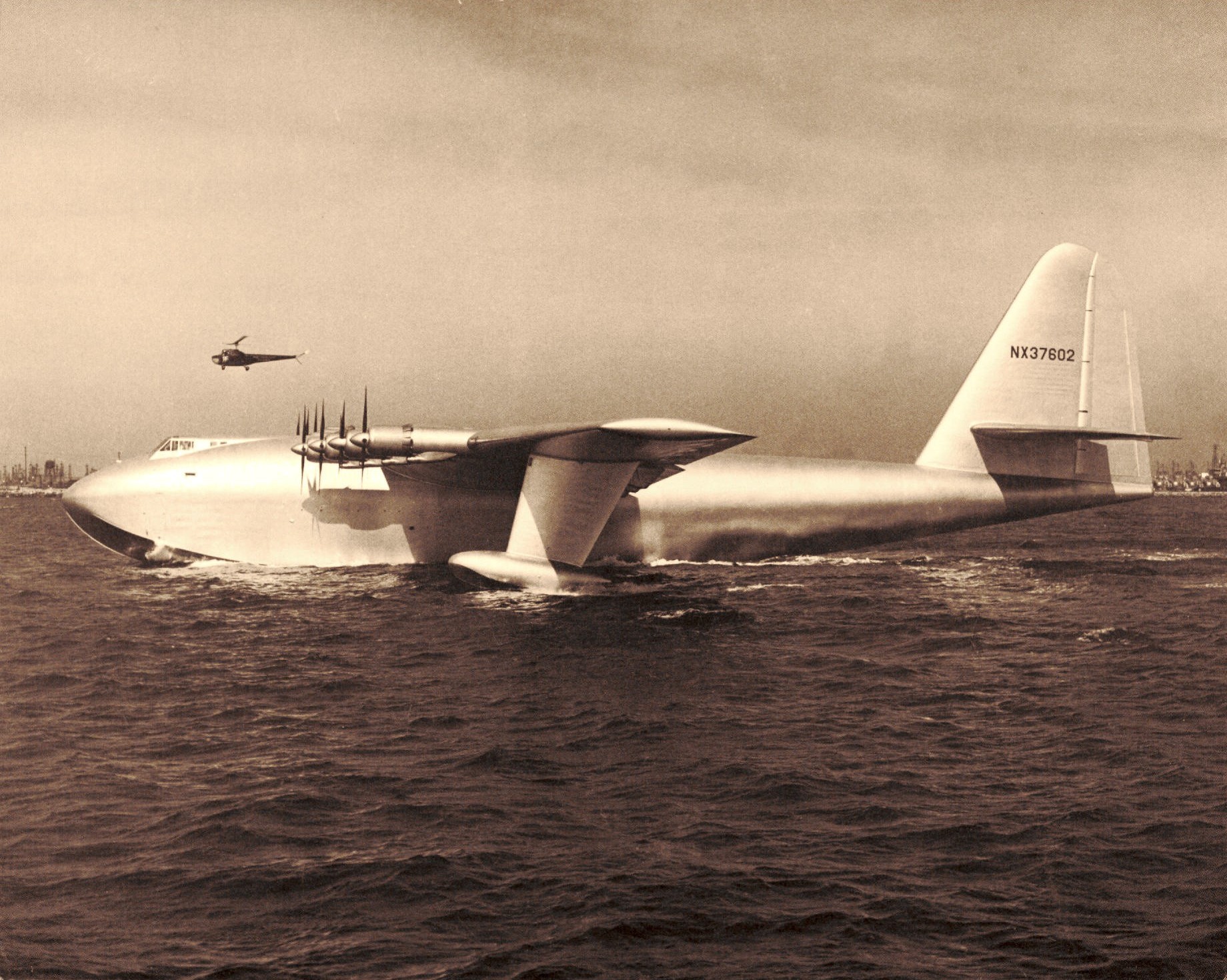 H-4 Hercules flying boat on the water, with an aircraft overhead.